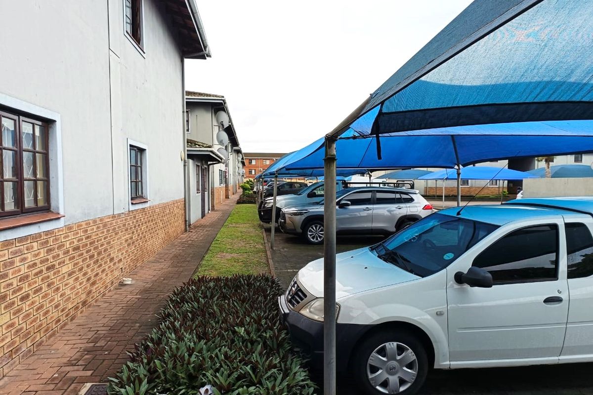 Exterior view of the apartment complex featuring a paved walkway, brick and plaster facade, and covered parking bays with shade cloth structures.