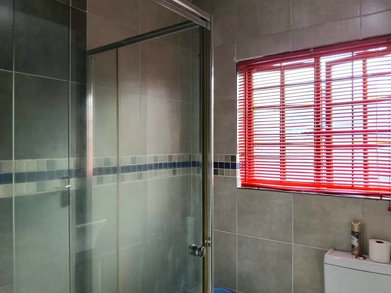 A functional bathroom featuring a glass shower enclosure, neutral ceramic wall tiling, and a window with red blinds.