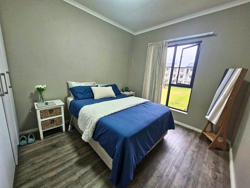 A well-maintained bedroom featuring modern laminate flooring, a built-in wardrobe, and an aluminium-framed window with views of the complex grounds.
