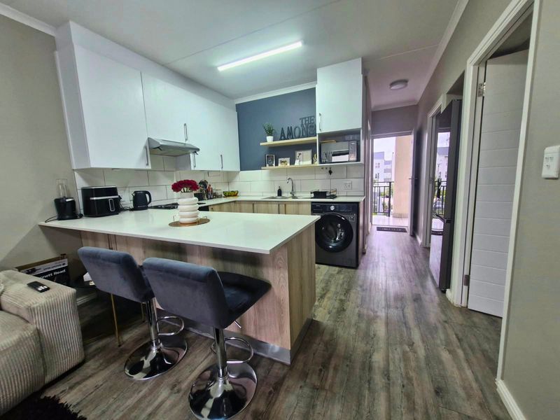 Modern open-plan kitchen featuring light wood cabinetry, stone countertops, a breakfast bar, and integrated laundry appliances, leading to a balcony.