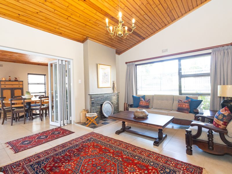 Spacious open-plan living area featuring a vaulted timber ceiling, a classic brick fireplace, and large windows, seamlessly connecting to the adjacent dining space.