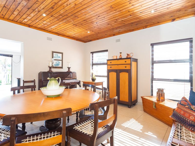 A bright dining room featuring a warm timber tongue and groove ceiling, tiled flooring, and large windows with venetian blinds.