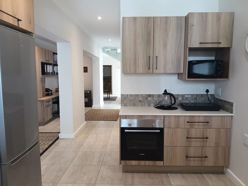 Modern kitchen area featuring light wood-grain cabinetry, stone countertops, mosaic backsplash, and integrated appliances, seamlessly flowing into an adjacent office workspace.