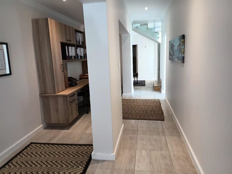 A bright office space featuring built-in wooden cabinetry and desk, leading into a hallway with a modern glass staircase.
