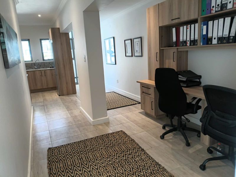 Modern office space featuring built-in wooden cabinetry and desk units, with an adjacent kitchenette area and neutral tiled flooring throughout.