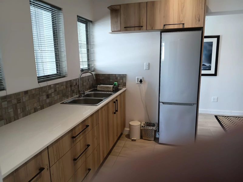 A compact, modern kitchen featuring light wood-grain cabinetry, stone countertops, a stainless steel double sink, and a tiled backsplash with natural light.