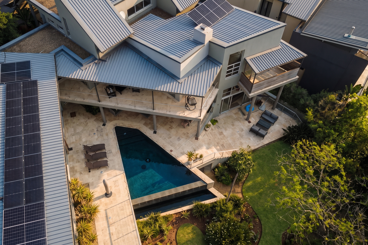 Aerial view of a modern residence featuring a metal roof with solar panels, a swimming pool, stone patio, and landscaped garden.