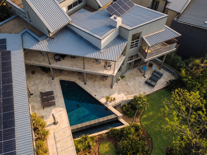 Aerial view of a modern residence featuring a metal roof with solar panels, a swimming pool, stone patio, and landscaped garden.