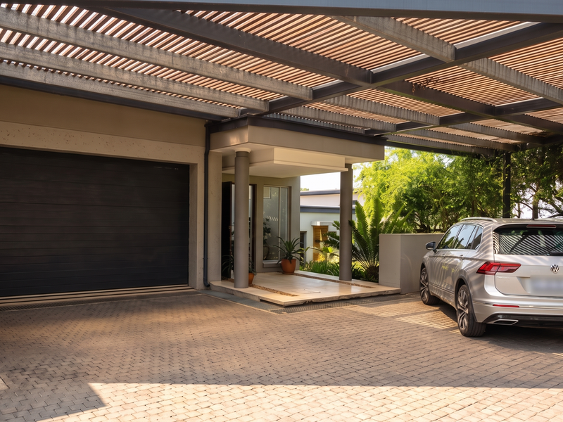 Modern front exterior featuring a spacious paved driveway, a large sectional garage door, and a stylish slatted pergola providing covered parking and entrance access.