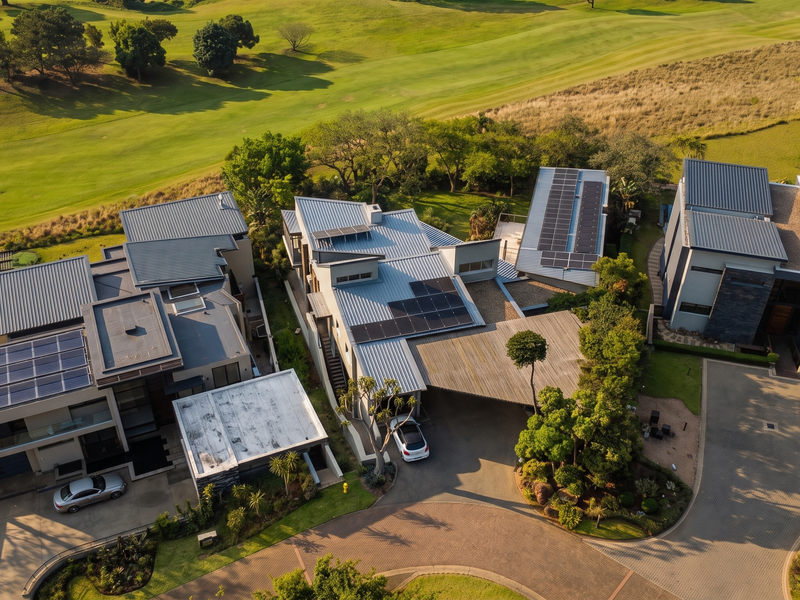 Aerial view of modern residential properties featuring metal roofs with integrated solar panels, paved driveways, and landscaped surroundings overlooking a golf course.