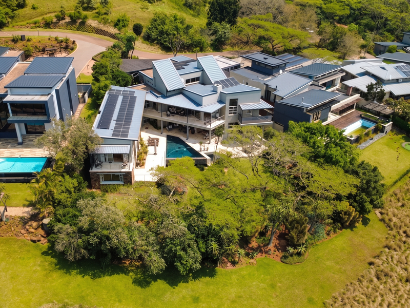An aerial view of a modern residential estate featuring multiple buildings with metal roofs, solar panels, swimming pools, and extensive landscaped grounds.