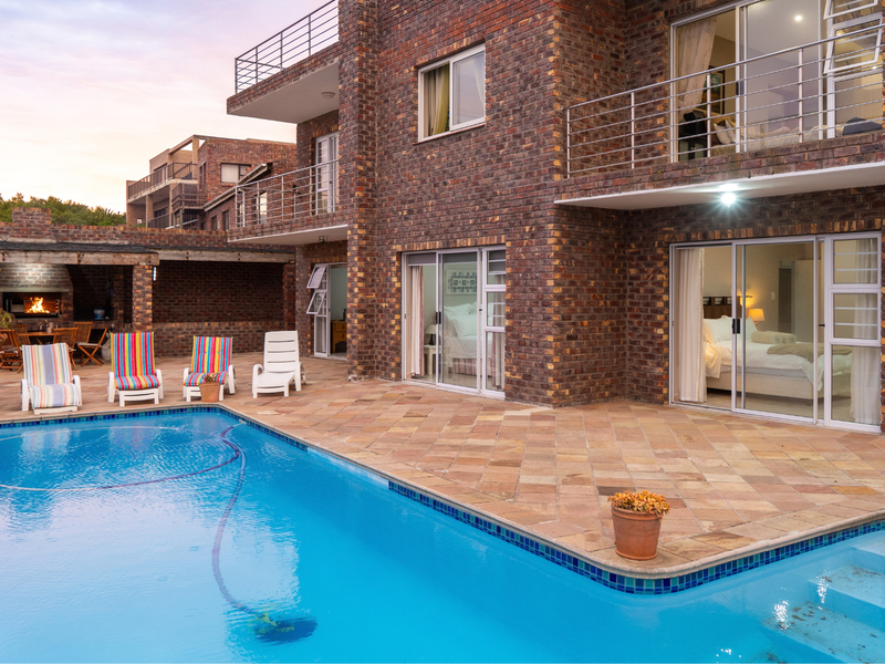 A spacious outdoor pool area featuring a stone patio, built-in braai, and direct access to the multi-story brick residence via sliding glass doors.