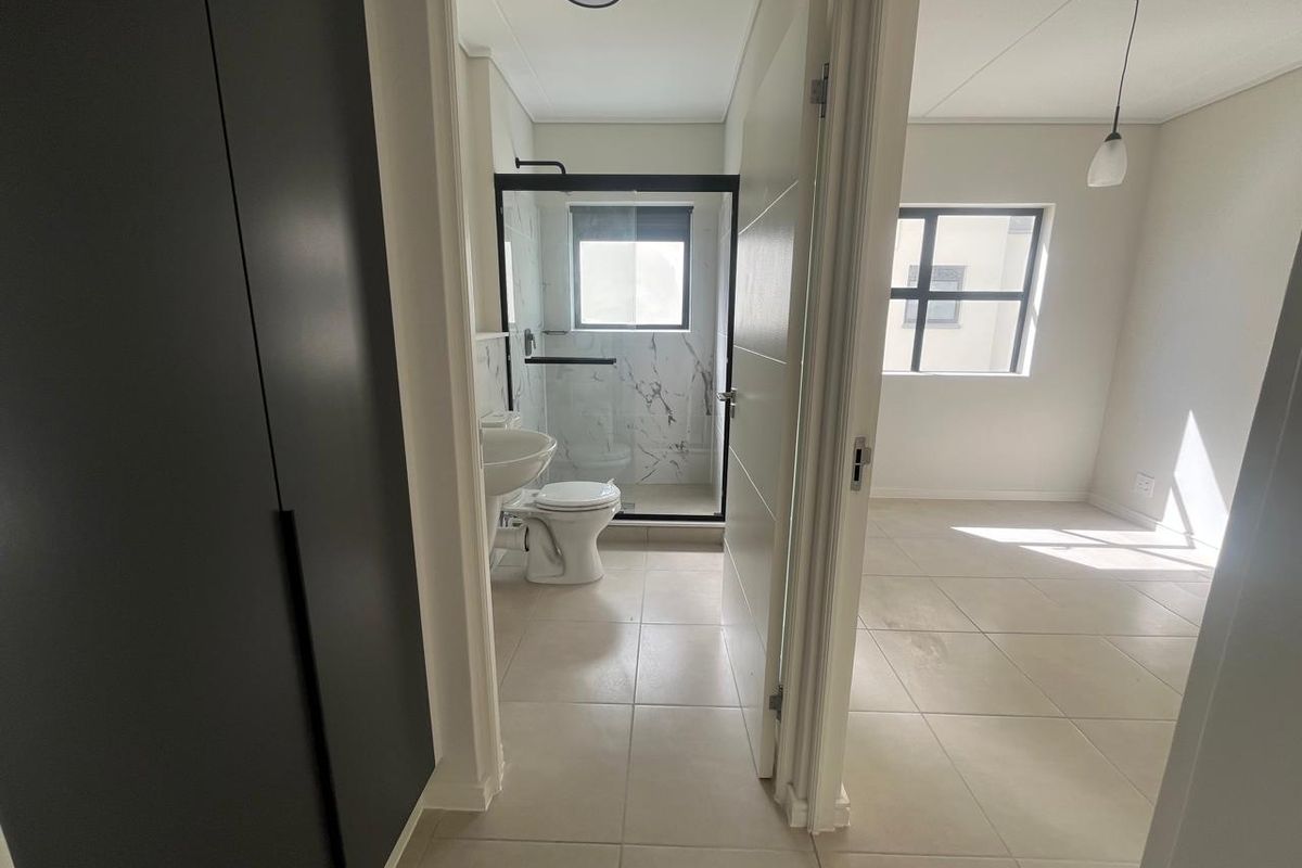 A modern, clean bathroom featuring a walk-in shower with black-framed glass, marble-look tiling, and a pedestal basin, adjacent to a bright, tiled living area.