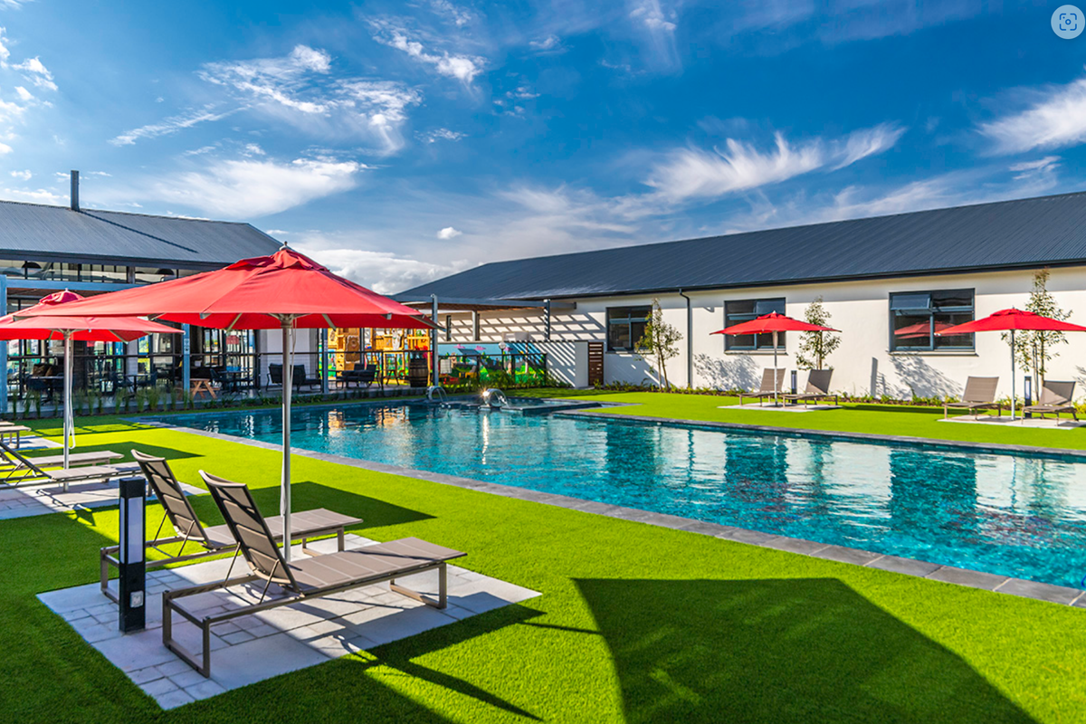 A well-maintained communal swimming pool area featuring artificial turf, paved surrounds, and modern building structures with metal roofing.