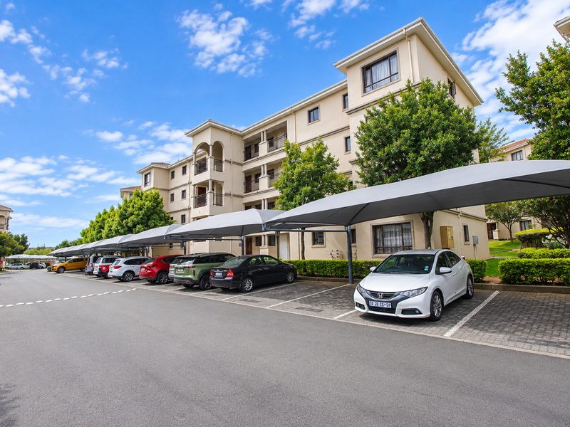 Exterior view of a multi-story apartment complex featuring paved parking bays with shade cloth carports and well-maintained landscaped garden areas.