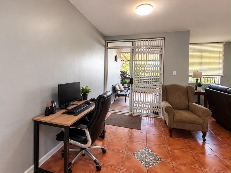A functional home office space featuring terracotta floor tiles, a sliding glass door with a security gate, and access to an outdoor patio area.