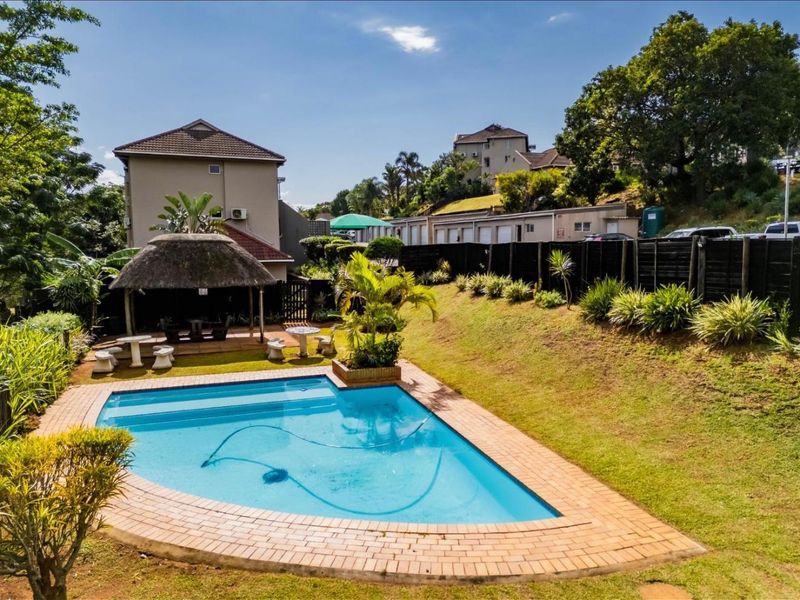 A well-maintained outdoor pool area featuring a swimming pool, brick paving, and a thatched gazebo set within a landscaped garden environment.