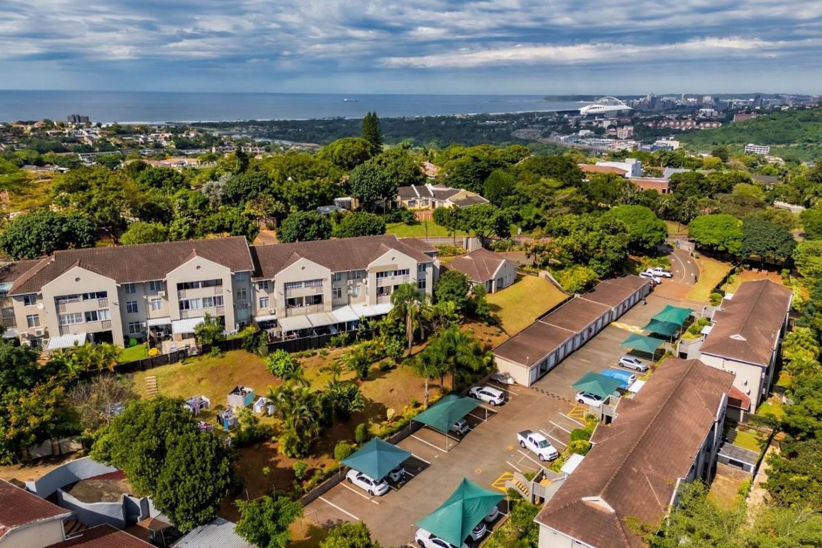 Aerial view of the apartment complex featuring tiled roof buildings, paved parking with shade cloth carports, and established communal garden areas.