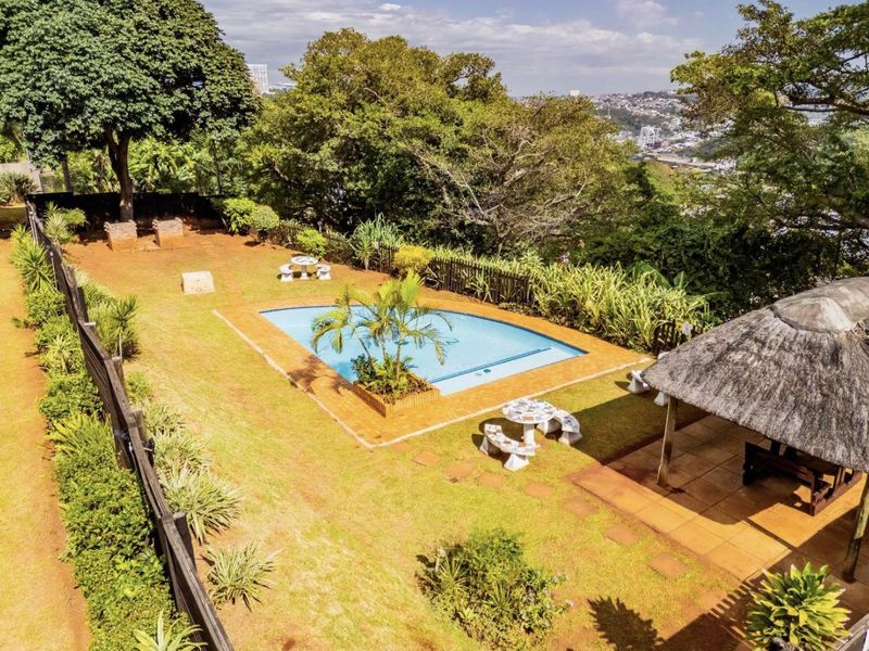 Outdoor pool area featuring a swimming pool, a thatched lapa, and a built-in braai, set within a spacious garden with scenic views.