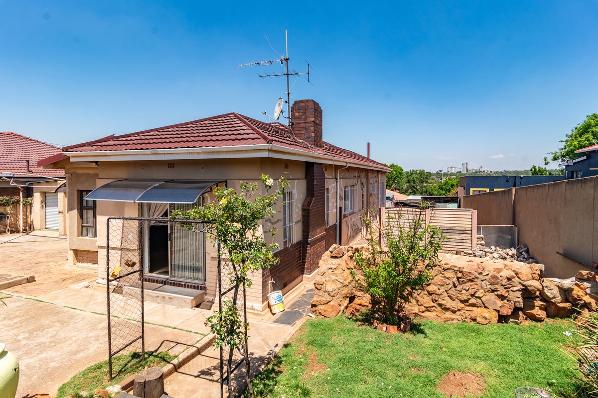 Rear view of a detached house featuring a tiled roof, brick chimney, paved yard area, and a stone retaining wall in the garden.
