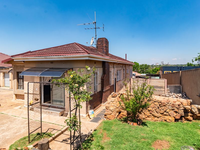 Rear view of a detached house featuring a tiled roof, brick chimney, paved yard area, and a stone retaining wall in the garden.