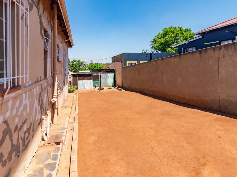 A view of the rear exterior area featuring a large open space, boundary wall, and side access to the main building structure.