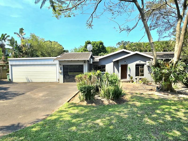 Front elevation of a single-story house featuring a double garage, asphalt driveway, and established garden surroundings.