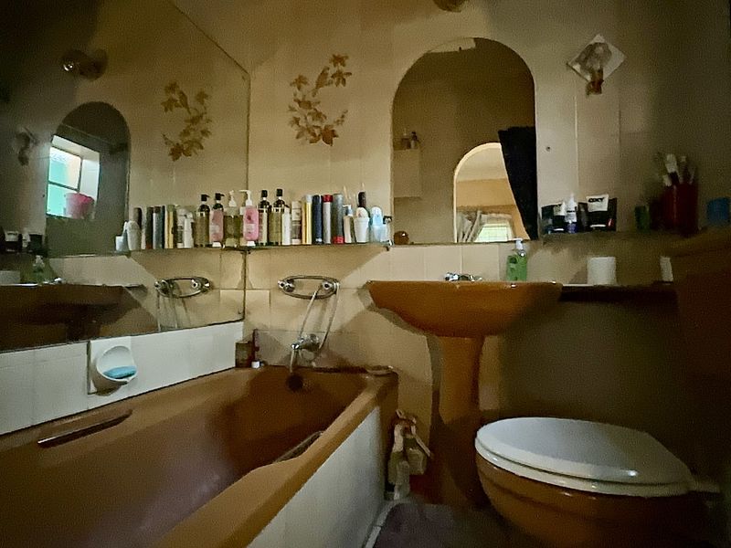 A dated bathroom featuring a pedestal basin, built-in bathtub, and toilet, with original wall tiling and a large wall-mounted mirror.