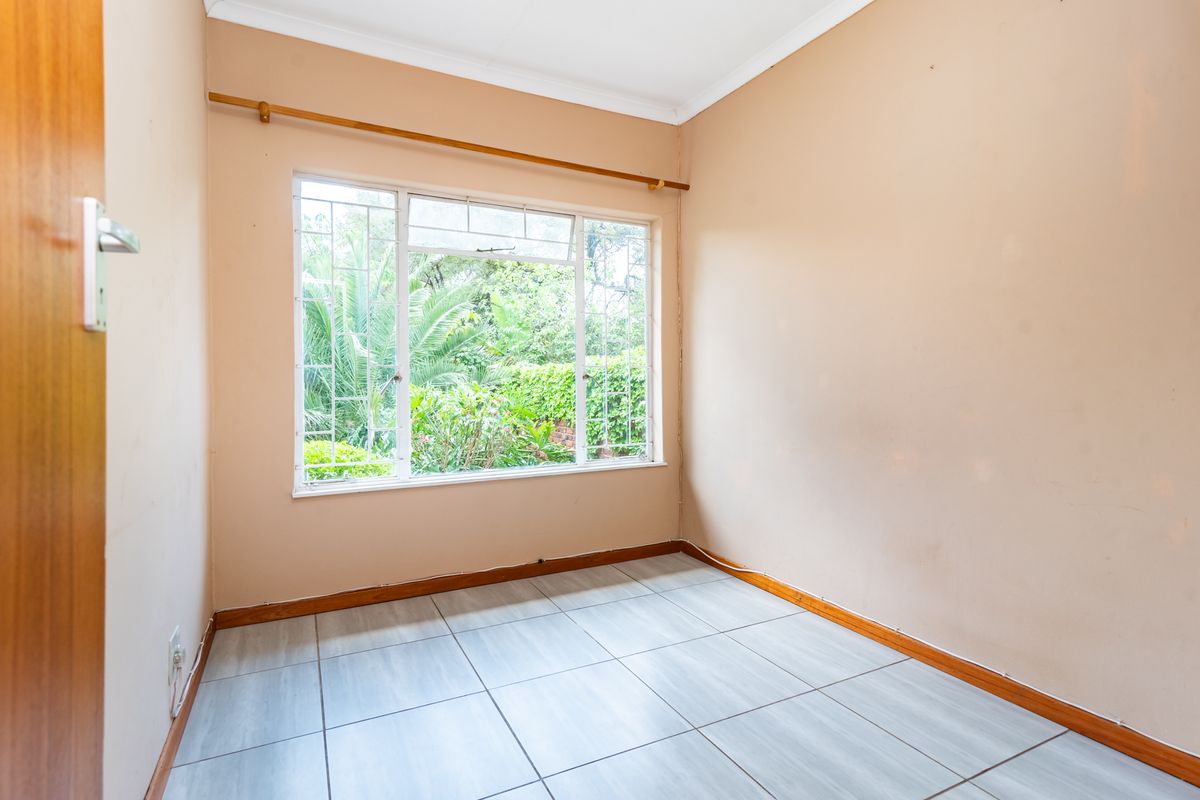 A vacant bedroom featuring light-colored tiled flooring, neutral wall tones, and a large window with security bars overlooking the garden.