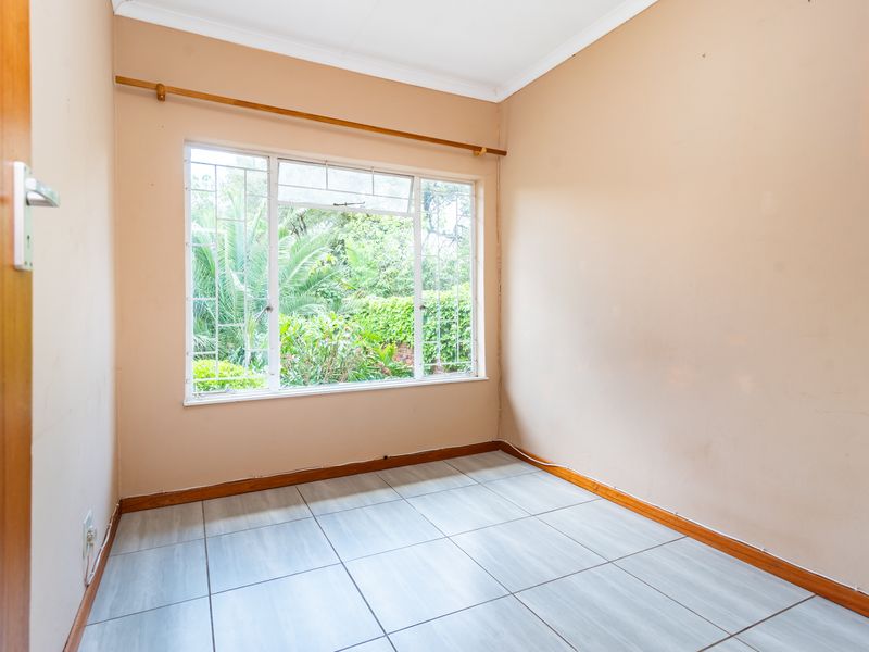 A vacant bedroom featuring light-colored tiled flooring, neutral wall tones, and a large window with security bars overlooking the garden.