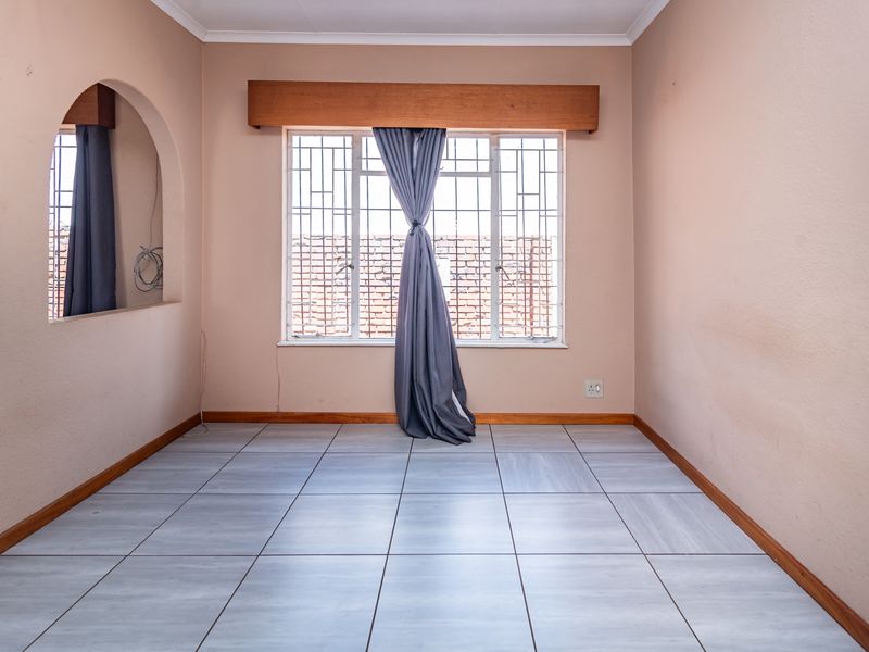 An empty living room featuring light-toned floor tiles, an arched architectural opening, and a window fitted with security bars and a wooden pelmet.