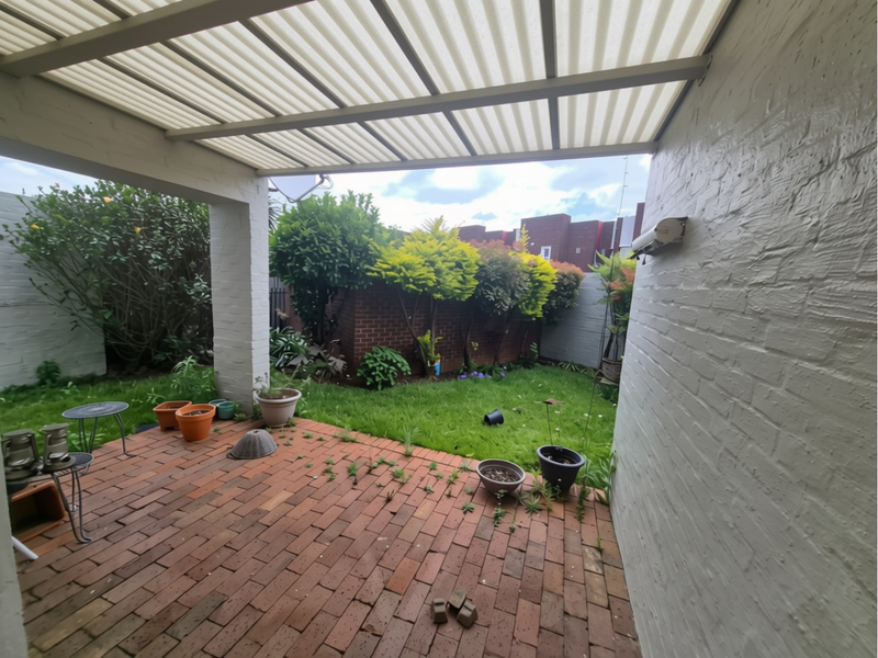 A covered patio area featuring brick paving and a translucent roof, overlooking a private garden space with established perimeter hedging.