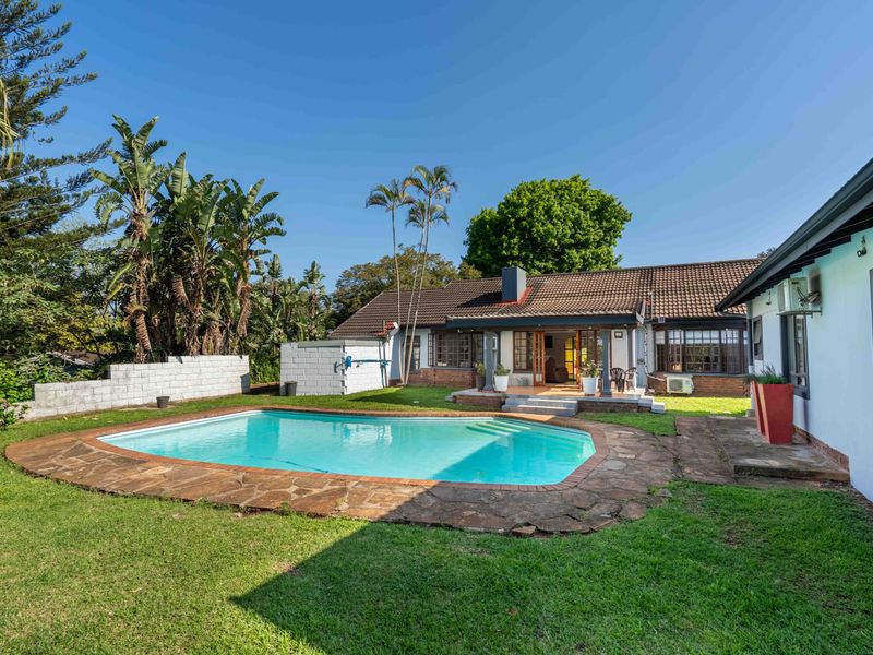 A spacious backyard featuring a swimming pool with stone coping, a paved patio area, and a well-maintained lawn set against the rear house elevation.