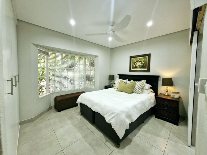 A well-maintained bedroom featuring a large bay window, neutral tiled flooring, recessed lighting, a ceiling fan, and built-in wardrobe storage.