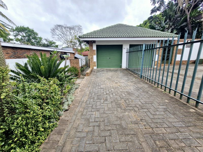 A double garage with green roller doors, featuring a brick-paved driveway and secure metal fencing in a well-established garden setting.
