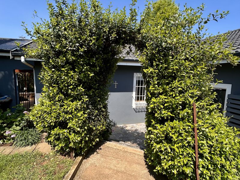 A rear exterior view featuring a paved walkway leading through a mature hedged archway toward the main dwelling with security-fitted windows.