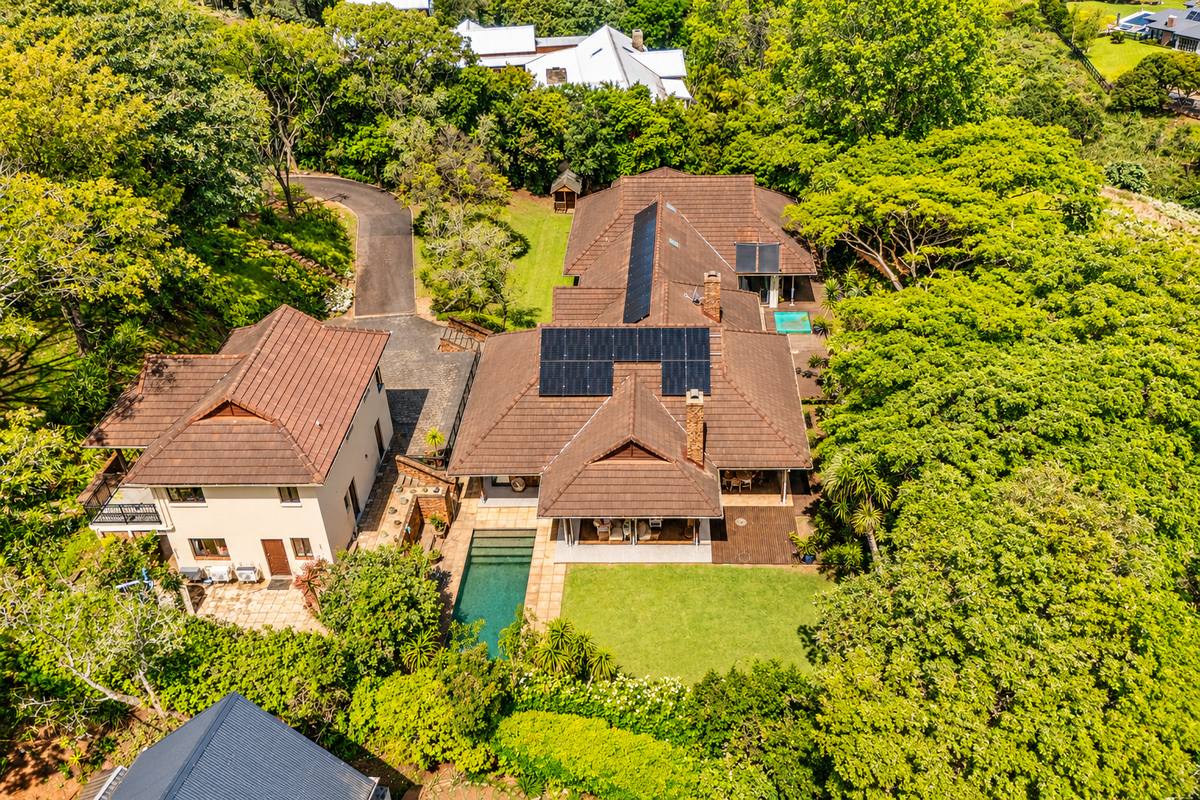 Aerial view of a residential property featuring a tiled roof with solar panels, a swimming pool, and extensive surrounding garden and mature trees.