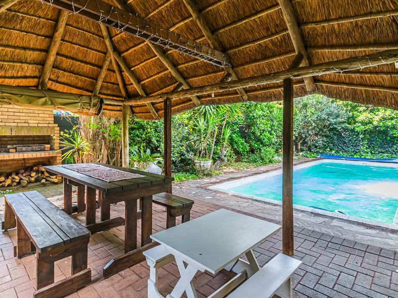 Covered outdoor patio featuring a traditional thatched roof, built-in brick braai, and direct access to a private swimming pool area.