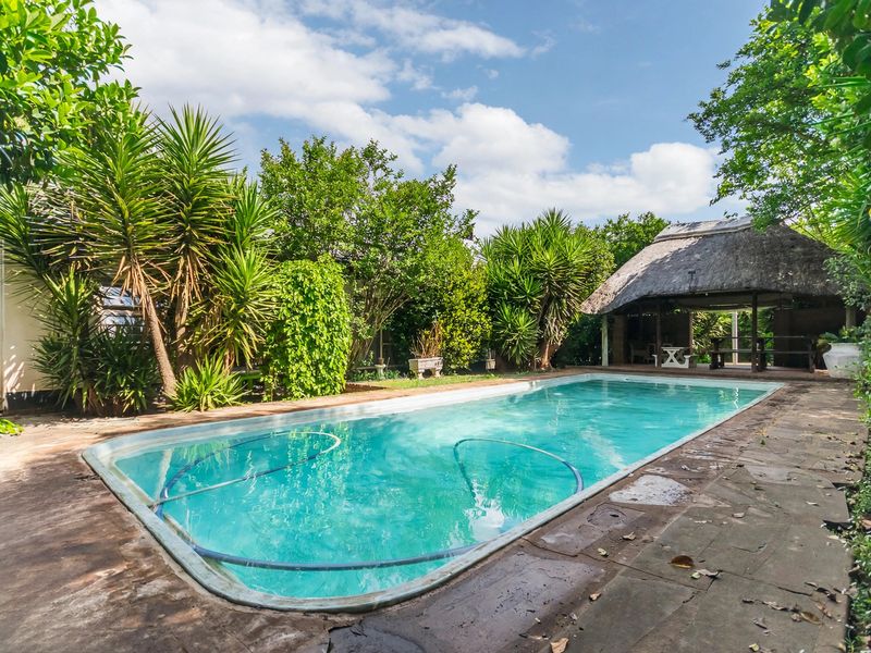Outdoor swimming pool area featuring a thatched lapa and stone paved surround, set within a lush, established garden.