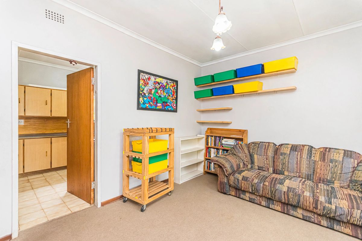 A functional bedroom featuring fitted wall shelving, neutral wall finishes, and carpet flooring, with an adjacent view into a kitchen area.