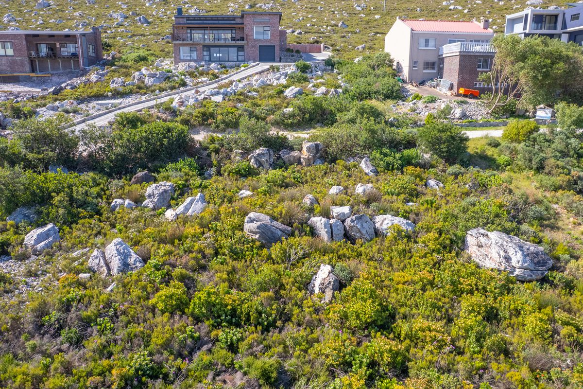 An aerial view of a sloped, undeveloped residential plot featuring natural rocky terrain and indigenous vegetation, situated in a hillside neighborhood.