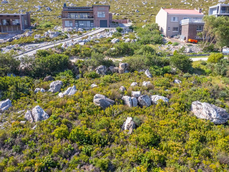 An aerial view of a sloped, undeveloped residential plot featuring natural rocky terrain and indigenous vegetation, situated in a hillside neighborhood.