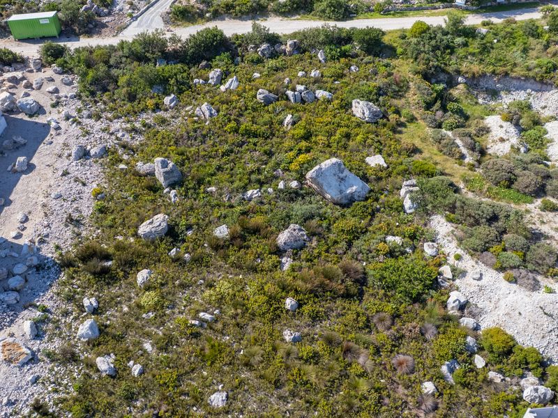 Aerial view of a vacant residential plot featuring natural rocky terrain and native vegetation, situated adjacent to an existing building structure.