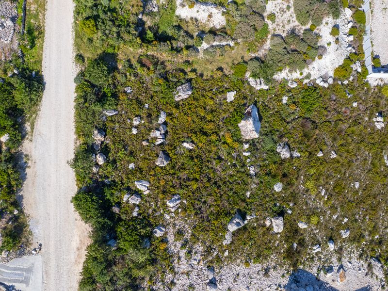 Aerial view of a vacant residential plot featuring natural vegetation, rock outcrops, and adjacent gravel road access.