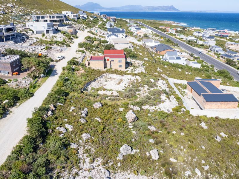 Aerial view of a vacant residential plot situated in a coastal development, featuring natural rocky terrain and proximity to established homes and the ocean.