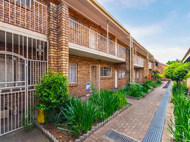 Exterior view of a face brick apartment complex featuring paved walkways, landscaped garden beds, and secure metal gate entrances.