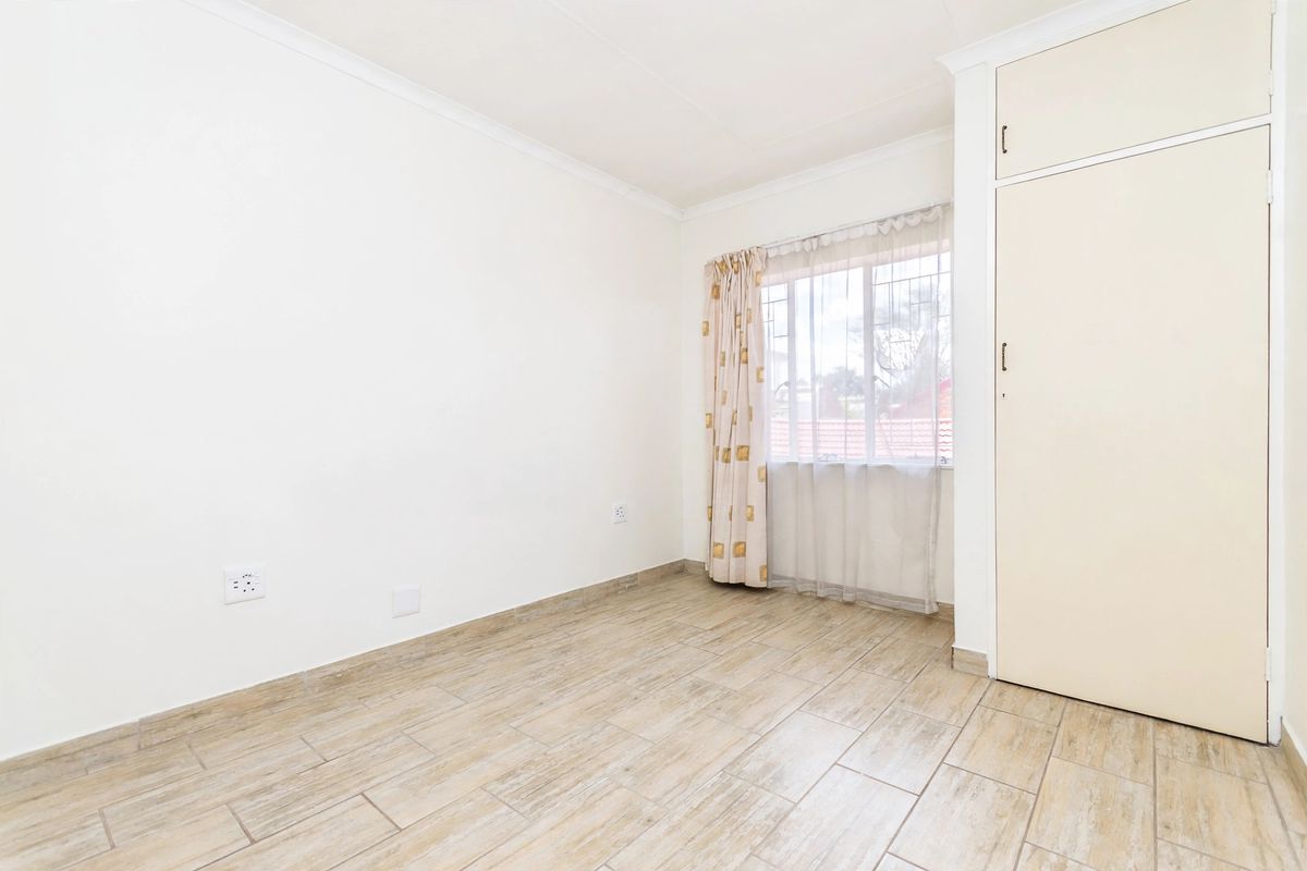 A bright, empty bedroom featuring neutral tiled flooring, a built-in wardrobe, and a window fitted with security bars.