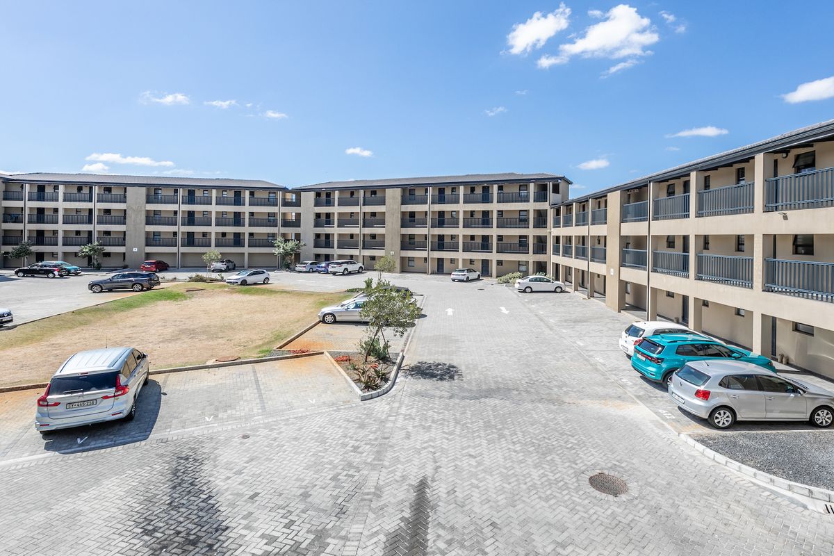 An elevated view of a multi-story apartment complex featuring a paved parking area, communal garden spaces, and private balconies for each unit.