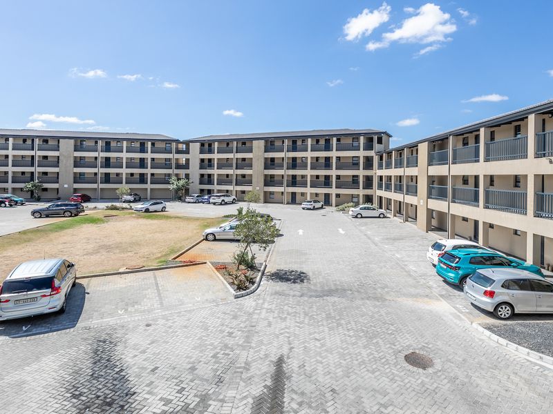 An elevated view of a multi-story apartment complex featuring a paved parking area, communal garden spaces, and private balconies for each unit.