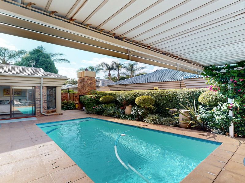 A well-maintained outdoor pool area featuring a covered patio, brick fireplace, and established garden landscaping, perfect for outdoor entertaining.